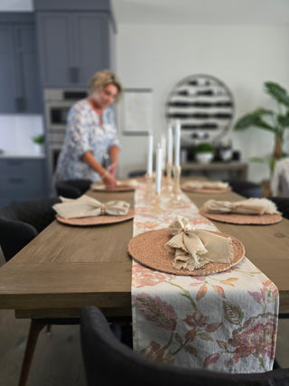 woman standing setting up a beautiful dining tabletop tablescape with a linen table runner in soft pink and coral colors.  Round woven placemats with fringed cream linen dinner napkins. Coral candle holders are on table with white taper candles.