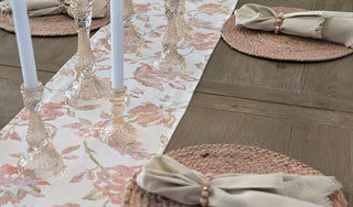 Table setting with floral linen table runner, coral glass candle holders, and round woven placemats with cream fringed linen dinner napkins with custom semi precious stone napkin rings