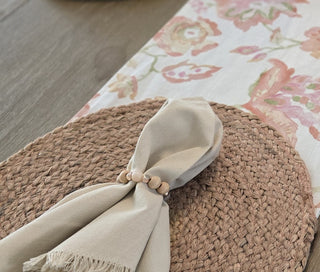Round woven placemat with a beige fringed linen dinner napkin with a custom rose gold hematite semi precious stone napkin ring around it on a floral linen table runner in soft pink and coral tones